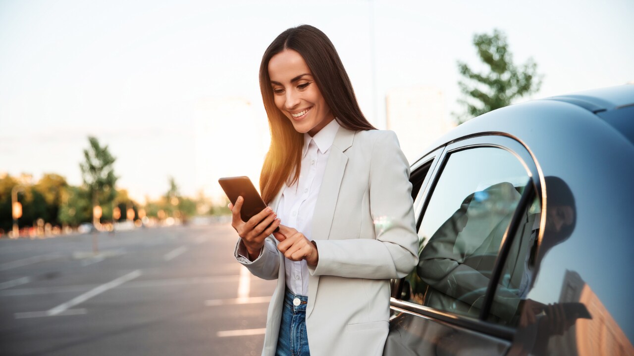 Woman smiling looking at her phone while standing outside of a car.