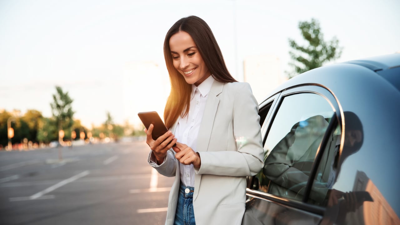 Femme souriante regardant son téléphone tout en se tenant à l’extérieur d’une voiture.