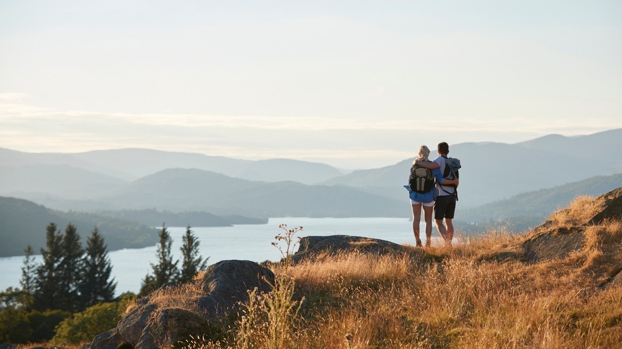 Rear View Of Couple Hugging On Top Of Hill On Hike Through Countryside In Lake District UK
